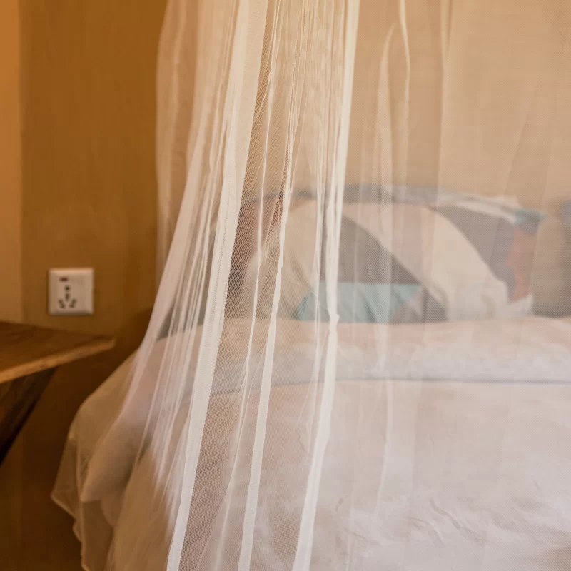 Close view on the bed with a white canopy style mosquito net. There is a power socket and a little wooden slap next to the bed