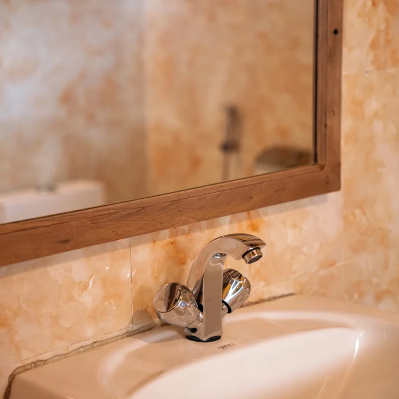 Bathroom sink above which is a large wooden framed mirror. The walls of the bathroom are covered with simple warm coloured tiles